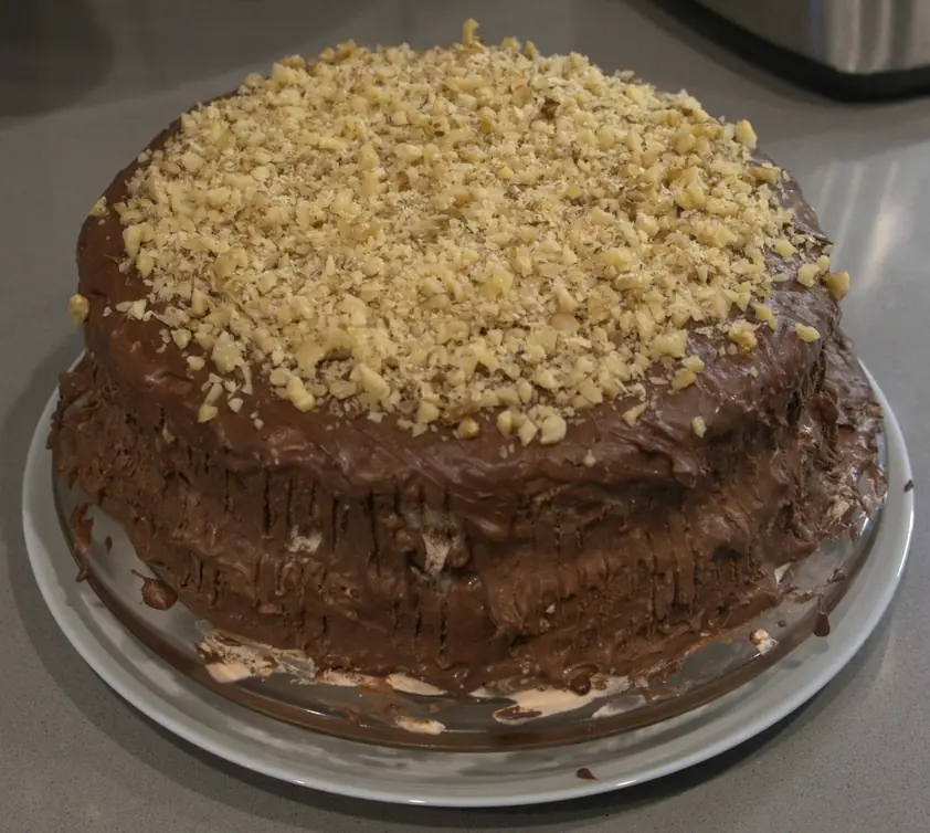 A cake coated in ganache with vertical lines down the side, and topped with chopped walnuts.